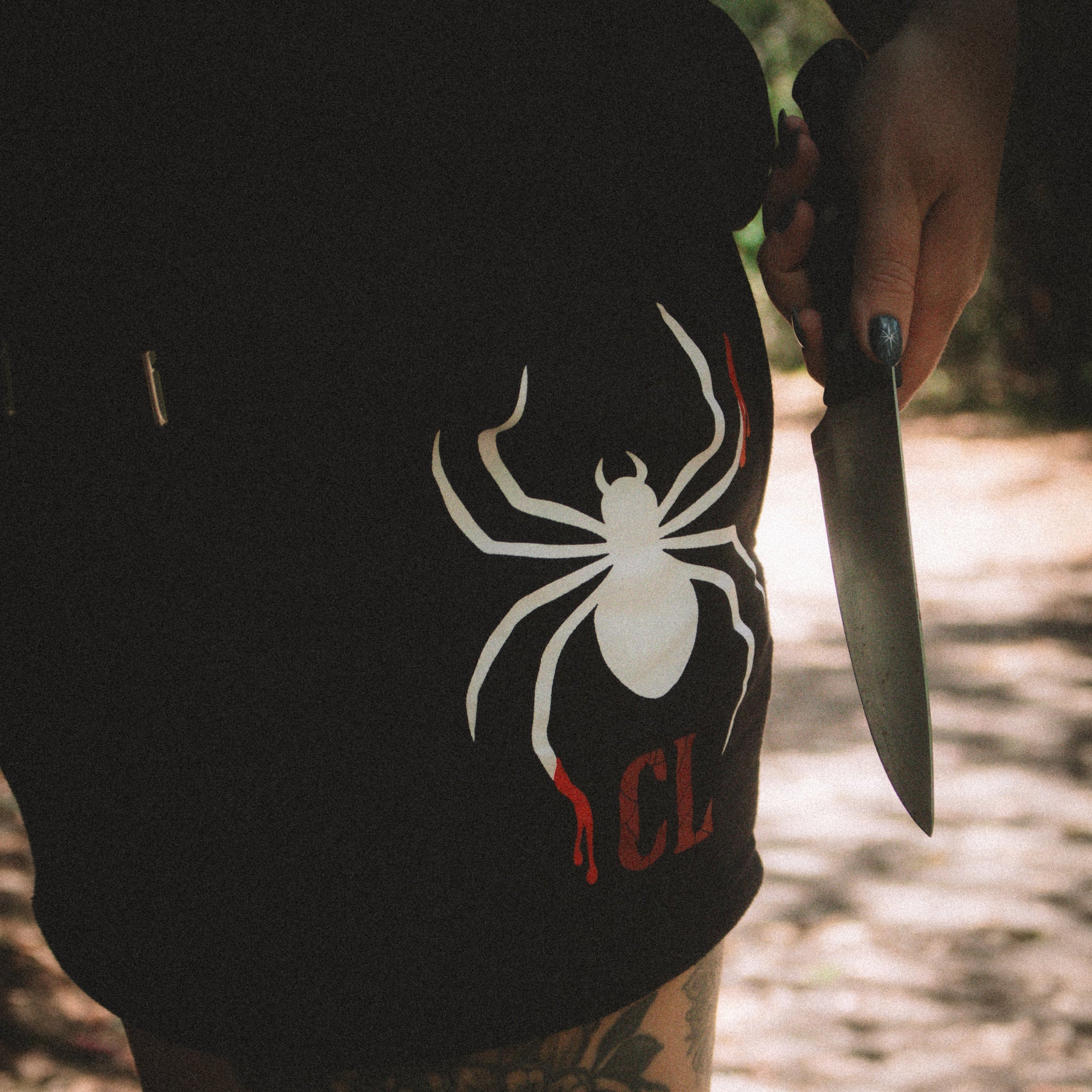 Person wearing a black shorts with a white spider logo, holding a knife in a forest setting.