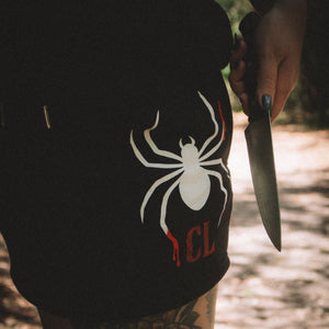 Person wearing a black shorts with a white spider logo, holding a knife in a forest setting.