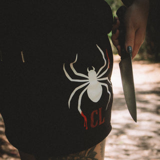 Person wearing a black shorts with a white spider logo, holding a knife in a forest setting.