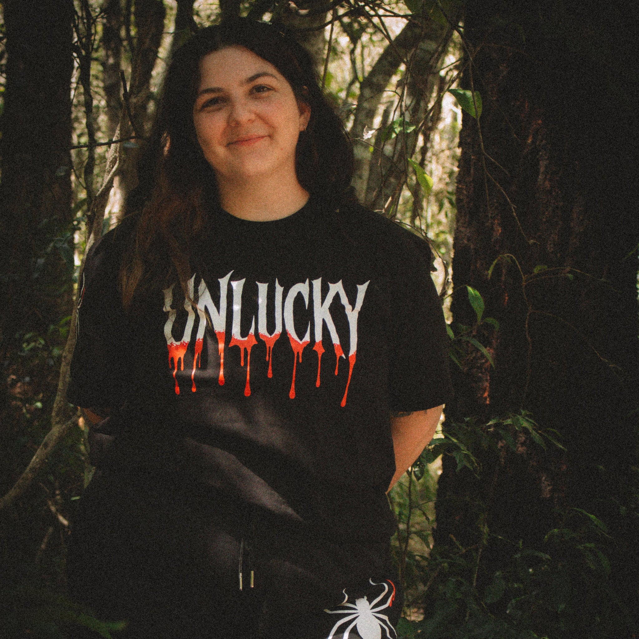 Person wearing a black t-shirt with 'UNLUCKY' design in a forest setting