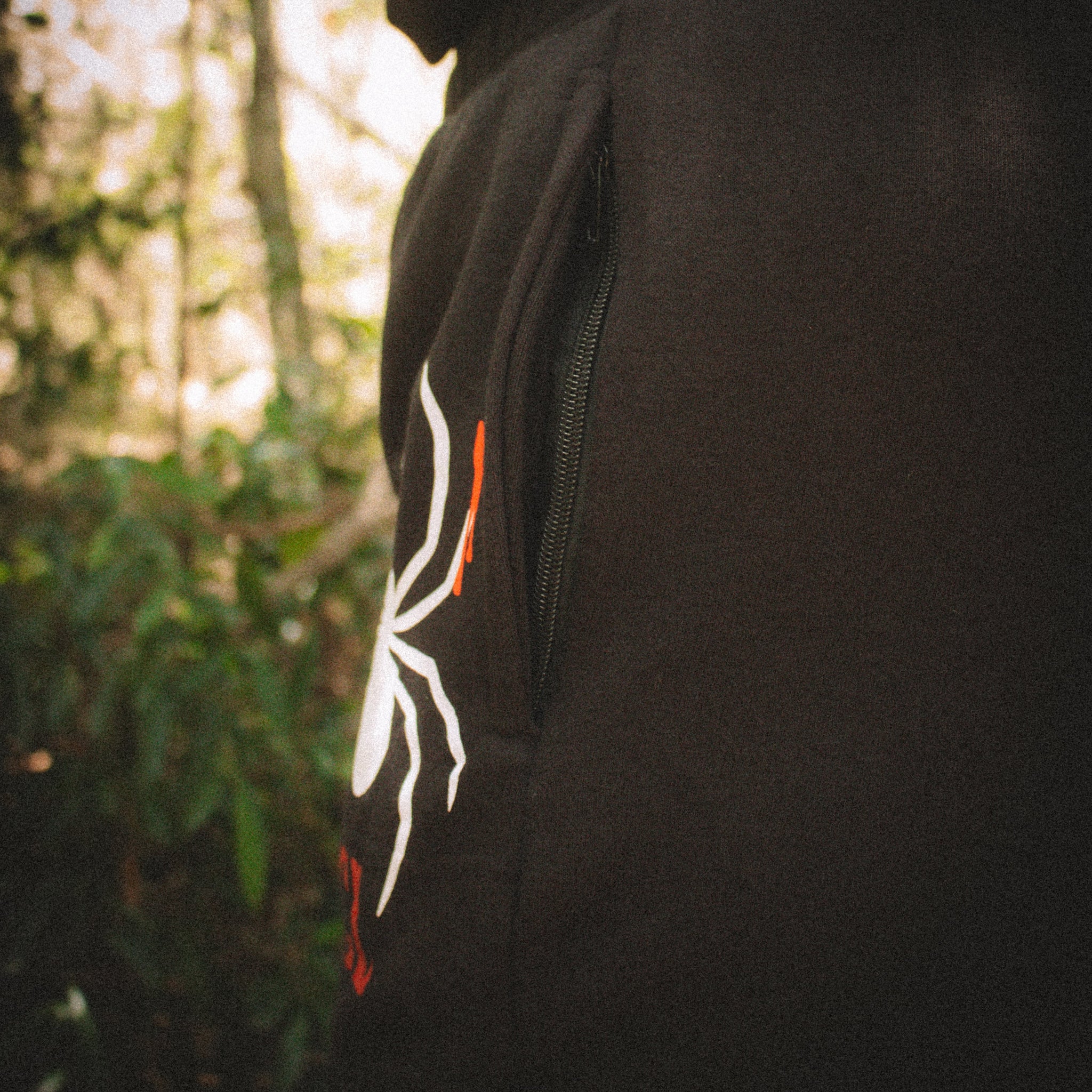 Black hoodie with a white and orange logo worn by a person in a forest setting