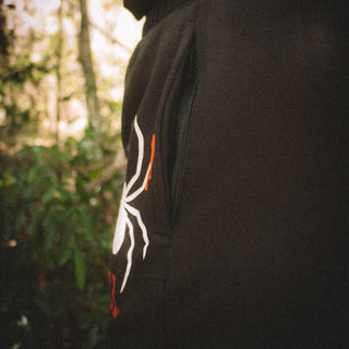 Black hoodie with a white and orange logo worn by a person in a forest setting