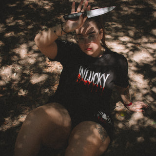 Person wearing a black t-shirt with 'WILUCKY' printed on it, sitting in a forest.