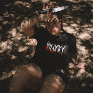 Person wearing a black t-shirt with 'WILUCKY' text, sitting on the ground with a knife.