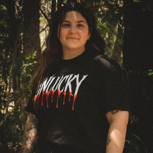 Person wearing a black t-shirt with 'UNLUCKY' text in a forest setting