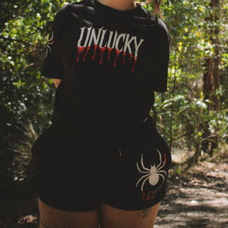 Person wearing a black t-shirt with 'UNLUCKY' text and spider design in a forest setting